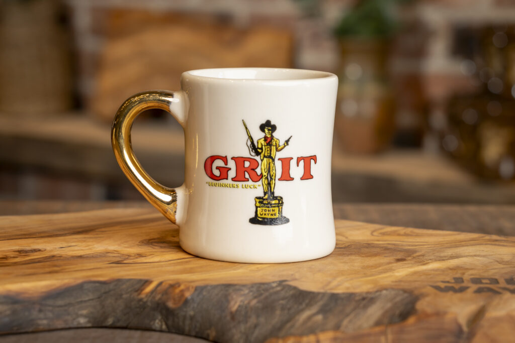 A John Wayne Gold Handle Mug featuring a gold handle and a cowboy design with the word 'GRIT.' Inspired by John Wayne’s tradition of gifting gold-handled mugs to cast and crew.